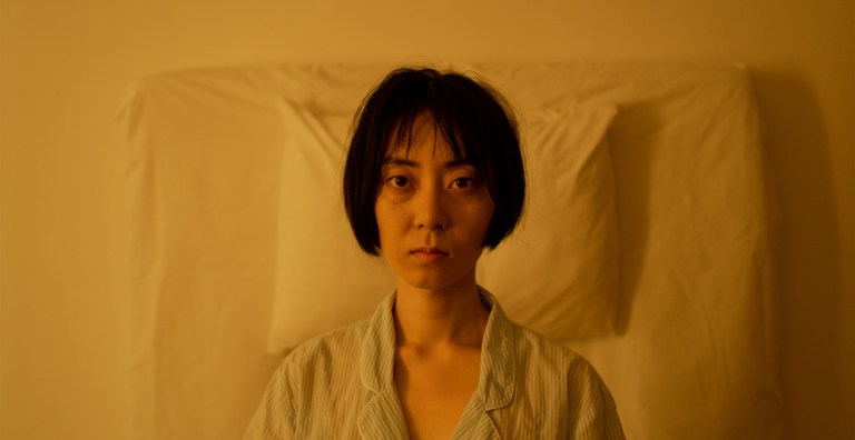 Portrait of a woman with short hair in striped pajamas, seated up against white pillows in a warm yellow-lit bedroom, facing the camera